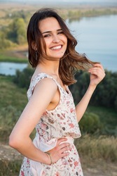 brunette woman with loose hair and perfect smile in dress with flower pattern stands on riverbank on windy summer day closeup, sunlight