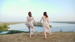 Graceful brunette barefoot women in long dresses walk joining hands along hilly riverbank at countryside on windy summer day backside view