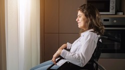 Young woman with organic damage of central nervous system sits in wheelchair looking out of window behind curtain at home, side view.