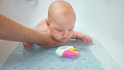 Father holds small daughter under belly in water. Baby girl plays with bright toy in bathtub. Child waves hands in water with amusement and curiosity