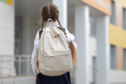 Long-haired girl with white backpack goes to modern school. Junior schoolgirl with long braids starts productive day at private primary school