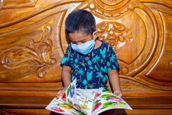 A baby boy is wearing a face mask and sitting on a bed reading a book. Facial hygienic mask for virus safety. Baby portrait in-home quarantine.