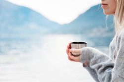 Woman with mug of coffee by winter sea, blue mountains. Cozy picnic with thermos of hot steamy beverage tea on beach. Girl is enjoying nature, life, relaxation, Christmas mood.