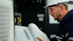 Caucasian production engineer in safety wear is reading the manual of a machine to find an error. A male factory worker is checking the industrial control panel of a robotic machine for maintenance.