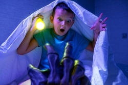 Scared boy with a flashlight hides under the blanket from the hand of a scary monster. Nightmare for children.