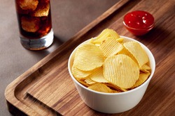 Potato chips in a bowl with dip sauce on wooden service tray and glass of coke. Close up view