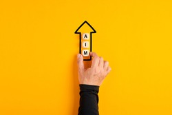 Male hand placing the wooden blocks with the word aim in an arrow pointing upwards on yellow background. Aiming high in business or education concept.
