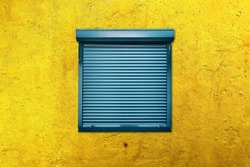 Square window with closed blue blinds or rolling shutter on a yellow weathered wall. Secrecy, mystery or protection concept.