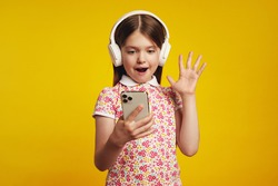 Surprised little kid in white headphones, holds modern smartphone and shows palm, happy to receive notification, wears summer clothes