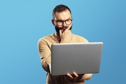 Portrait of a doubtful bearded man holding laptop computer isolated over blue background 