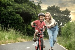 Mother teaching son to ride bicycle. Happy cute boy in helmet learn to riding a bike on the bike path in summer raining day at sunset time. Family weekend.