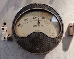 a large round vintage industrial ammeter with an analogue dial with numbers with standard electrical symbols on a white dial on a grey background