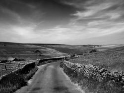 monochrome image of a narrow country lane surrounded by dy stone walls and dramatic clouds in a sunlit rural pennine landscape on the old howarth road in calderdale west yorkshire
