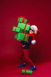 little boy in dark glasses and a Santa hat holds a pyramid of Christmas gift boxes on a red background in the Studio. the child smiles with pleasure and gets a lot of gifts for Christmas. Advertizing.