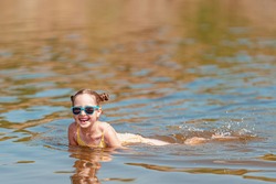 Portrait of a happy little girl in sunglasses lying in the water on a sunny summer day. A child enjoys a summer children's holiday on the lake shore. Active holidays. Vacation on the sea or river