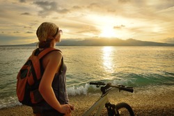 Young woman with backpack standing on the shore near his bike and enjoying the sunset over the sea on the background of the island Negros, Philippines.