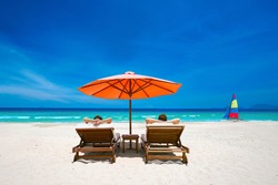 Couple on a tropical beach relax in the sun on deck chairs under a red umbrella.  Travel  background .
