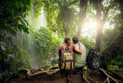Couple travelers with backpacks relaxing in greens jungle and enjoying view in waterfall. Island Lombok, Indonesia.Traveling along mountains and rain forest, freedom and active lifestyle concept
