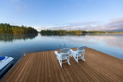 Beautiful morning landscape panorama, under a blue sky, of a lake in cottage country in Ontario, Canada. Two white Adirondack chairs facing the calm water are sitting on a new wooden dock.