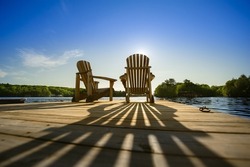 Cottage life - Sunrise on two empty Adirondack chairs sitting on a dock on a lake in Muskoka, Ontario Canada. The sun rays create long shadows on the wooden pier.