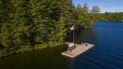 Two brown Adirondack chairs on a wooden pier facing a lake in Muskoka. Across the calm water is a brown cottage nestled among green trees. Canada flag is waving on a pole.