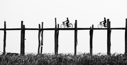 Black and white photo of old wooden bridge and silhouettes of two girls riding bikes , U Bein Bridge is a crossing that spans the Taungthaman Lake near Amarapura, Myanmar