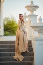 A beautiful young woman in a historic eighteenth century gold dress stands on the stairs of the mansion. Princess in the palace. fabulous image