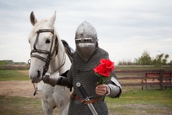A knight with a white horse and flowers in his hand. A romantic image. A young guy on a horse in medieval armor. Fantasy. Gives roses