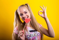 beautiful blond millennial with pink lollipop biting and sucking posing against poppy yellow background making an expression of how tasty and delicious this heart shaped candy thing is. text area 4 u
