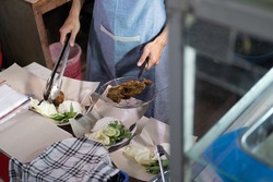 close up of the cook's hand holding the drain utensil and tongs while preparing side dishes and rice packaged orders for customers at the stall