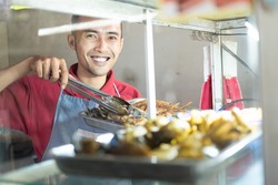 The smiling Asian male waiter takes the fried chicken on the iron tray in the food stall display window