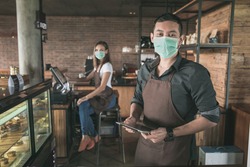 cafe owner wear face mask at his coffee shop using tablet pc. his partner sitting in a background. open for business in new normal protocol