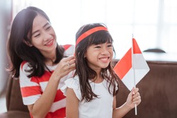 mother and daughter celebrating indonesian independence day at home wearing red and white with indonesia flag. mom tying ribbon on her daughter's forehead