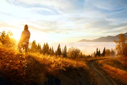 young woman hiking at mountain peak above clouds and fog Hiker girl wrapping in warm poncho outdoor.Early morning.Misty mountain.Young girl over the clouds in the valley looking at calm autumn sunrise