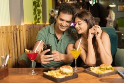 Indian Couple Looking in smartphone while enjoying food and drink at restaurant.