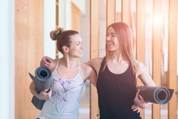 Two happy sporty women friends hugging, holding yoga mat and ready to start workout together yoga or pilates exercise indoor. Sun glare effect.