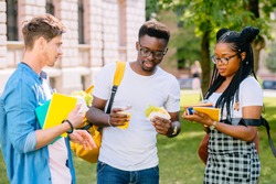 Group of three serious multiethnic students people coworkers talking, sharing ideas during lunch break standing outdoor university campus with books and backpacks. University education concept