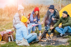  Group friends camping leisure and destination travel. Five people sitting around camp fire drinking hot tea, warming, joking together. Tourism relax and travel near lake in holiday. Sun glare
