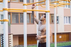 Young topless muscular athlete hanging on sports bar while pulling up on sportsground during workout.Naked torso man looking away, relaxing after exercises. Sport, lifestyle and people concept.