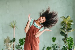 Eco friendly mixed race playful girl gardener fooling around shaking head with floating hair. African american woman in brown overalls and strip t-shirt on green background. Zero waste concept.