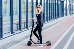 Full height photo of blond woman in black eyeglasses and wearing black clothes riding electric scooter on empty city street.Eco-friendly transport. Safety concept for road users. Rent scooter.