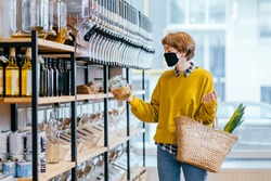 Shopping, food, sale, consumerism concept. Blond short haired woman in face protective mask holds glass jar with soya, buying at grocery store Reusable wicker basket for shopping. Zero waste concept.