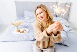 Portrait of a wonderful young blond woman in cozy sweater enjoying holding a sparkling sticks at home in winter time. People, holidays, celebrating, party, emotion and glamour concept.