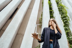Wide angle bottom view of businesswoman or female student dressed in a business suit with wind turbine model having conversation with smart phone outdoors. Renewable energy concept.