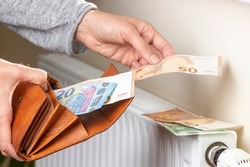Woman's hand takes euro money banknotes from wallet and places on heating radiator battery with temperature regulator. Expensive heating costs during cold season. Paying bills
