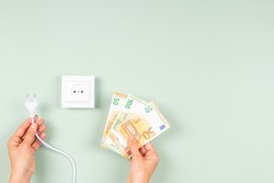 Woman hands holding electric power plug and Euro banknotes near white electric socket on light green background. Electricity cost and expensive energy concept