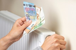 Woman hand holding euro banknotes and adjusting temperature of central heating radiator at home. Family pay money for home heating. Expensive heating costs