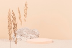 Round beige platform podium and grungy concrete stone on white beach sand with dry bent plant in foreground. Minimal creative composition background for cosmetics or products presentation. Front view