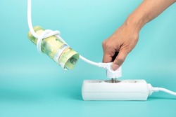 Woman hand holding electric power plug with Euro banknote on cord and plugging it in power strip extension cord on blue background. Electricity cost, electric prices increase, expensive energy concept