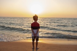 Cute toddler Asian boy standing alone on the beach at sunset. Child standing on the beach near sea and looking on the waves, silhouette of the boy on the seaside in sunset.